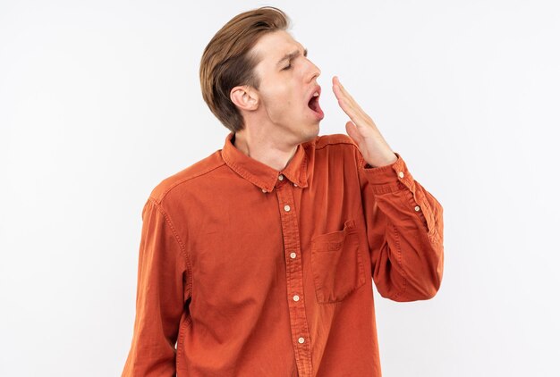 Yawning young handsome guy wearing red shirt covered mouth with hand isolated on white wall