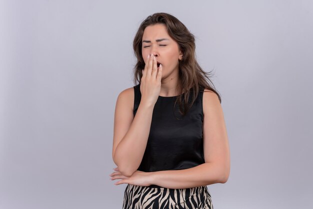 Yawns caucasian young girl wearing black undershirt put her hand on mouth on white background