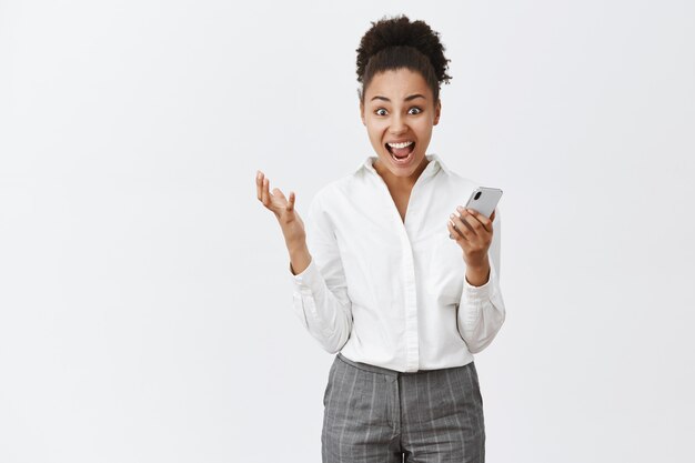 Yeah girl I did it. Portrait of shocked and surprised happy creative businesswoman with dark skin and bun hairstyle, gesturing while holding smartphone, reading impressive good news over grey wall