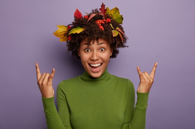 Yeah, its cool! Overjoyed woman with Afro hair decorated by colorful leaves, enjoys autumn time, raises hands and shows rock n roll gesture, dressed casually, feels energized isolated over violet wall