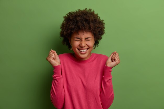 Yeah, this is my opportuity. Joyful Afro American woman rejoices success, clenches fists with triumph, has wish to win, celebrates achievement, wears rosy sweater, isolated on green wall