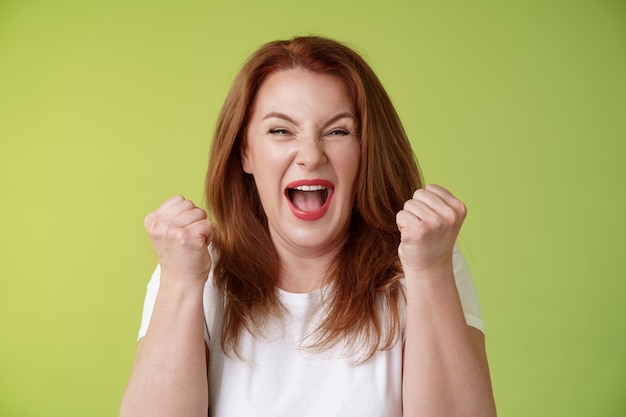 Yeah we did it joyful lucky redhead middleaged female winner pump fists up celebration success gesture yelling triumph joy smiling broadly celebrate awesome news stand green wall happy