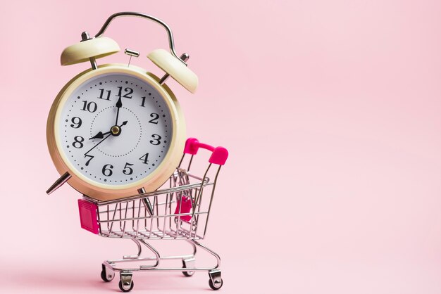 Yellow alarm clock inside the miniature shopping trolley against pink background