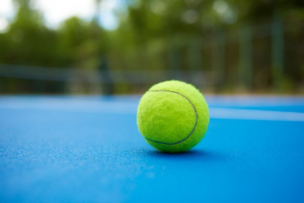 Yellow ball is laying on blue tennis court carpet. Blurred green plantings and trees behind.