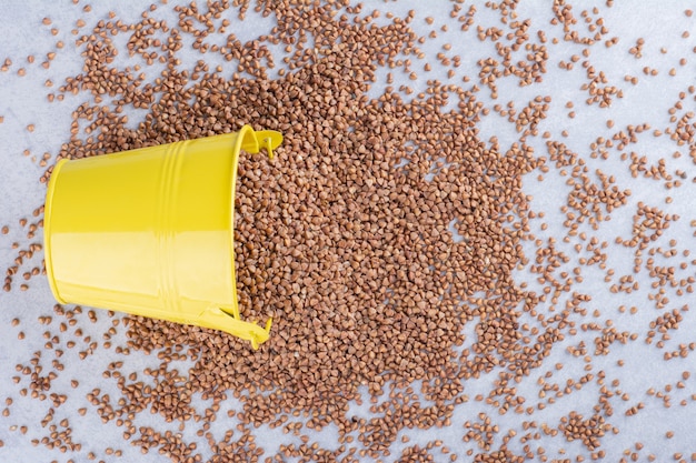 Yellow bucket lodged in a pile of scattered buckwheat on marble surface