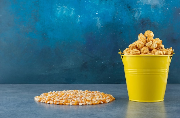 Желтое ведро конфет попкорна рядом с аккуратной грудой кукурузных зерен на синем