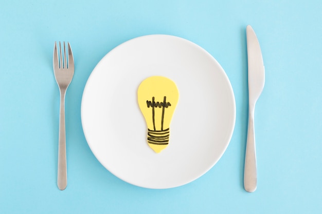 Yellow cutout light bulb on white plate with fork and butter knife against blue background