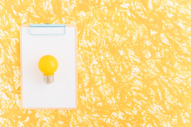 Yellow light bulb on white paper over clipboard against yellow backdrop