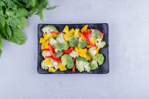 Yellow and red bell pepper and broccoli salad on a platter next to a pile of greens on marble background. High quality photo