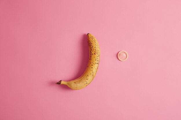 Yellow ripe banana and condom on pink background. Contraception and safety.