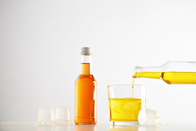 Yellow tasty lemonade drink pours from bottle to glass with ice cubes near closed sealed unlabeled bottle with orange beverage