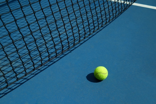 Желтый теннисный мяч лежит возле черной открытой сетки теннисного корта.