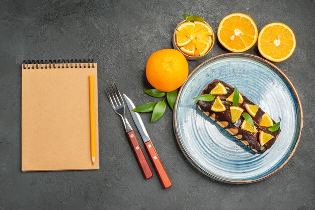 Yellow whole and cut lemons tasty cakes with fork and knife next to notebook on dark table