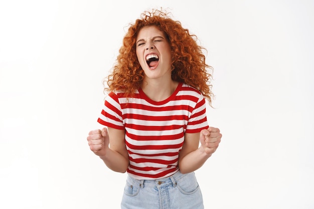 Yes finally success Attractive curly redhead wild girl ginger hairstyle close eyes shout joyful enjoy victory winning awesome prize achieve goal clench fists triumphing celebrating white background