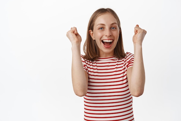 Yes hooray. Happy blond girl cheering, raising hands up and scream yes, smiling broadly, celebrating victory, achieve goal or win prize, triumphing, white background