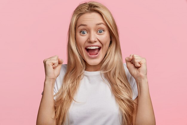 Yes, I done it! Excited cheerful female model with joyful expression, cheers and clenches fists, celebrates her success, has appealing look, poses against pink wall Achievement