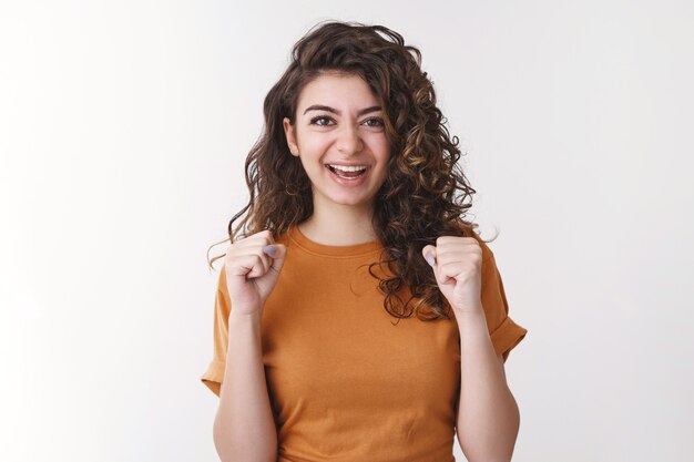 Yes lucky me. Happy triumphing young joyful armenian woman curly-haired clench fists celebrating successful win lottery smiling say yeah, excited good positive result, standing white background