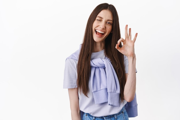 Yes Okay Cheerful brunette woman winking smiling and showing ok gesture approve and agree like something praise nice work perfect choice standing over white background