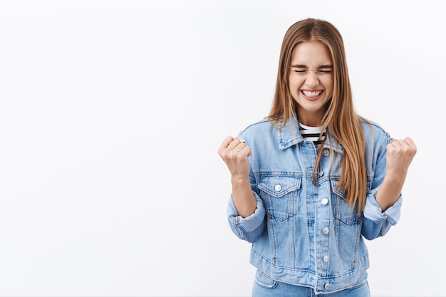 Sì, l'ha fatto. ragazza bionda felice e allegra che si rallegra per la vittoria, vince il premio, raggiunge l'obiettivo, pompa a pugno con gli occhi chiusi e il sorriso ottimista, guadagna l'obiettivo, celebra il successo, trionfa sul muro bianco