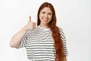 Free photo yes. smiling redhead female model showing thumbs up. girl with red long hairstyle and freckles likes smth good, give approval, agree or praise you, standing over white background