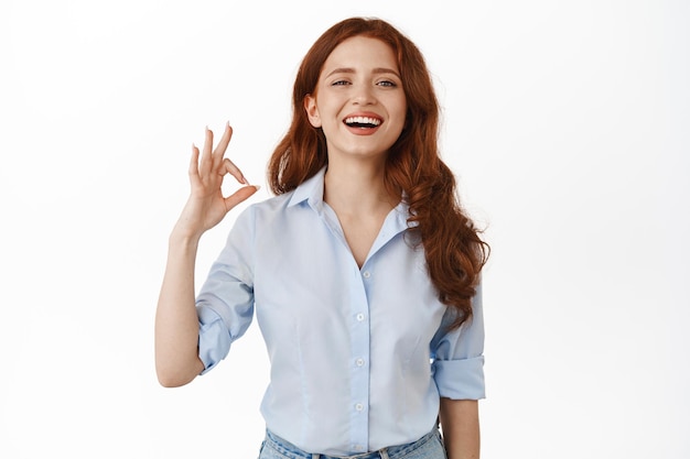 Yes, very good. Happy smiling business woman with red curly hair, nod pleased, show okay OK gesture and laughing, approve and like something nice, praise great job, white background