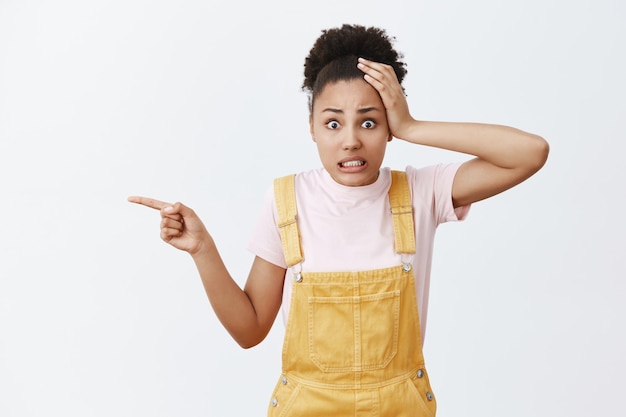 Yikes, what is that. Portrait of shocked worried and confused attractive african-american female with curly hair in fashionable yellow overalls, holding hand on head, pointing left with index finger