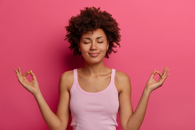 Free photo yoga and meditation concept. relaxed satisfied dark skinned woman holds hands in mudra gesture, feels peaceful after hard day, keeps eyes closed, controls her feelings, stands in lotus pose.