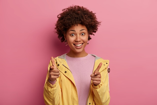 You are chosen. Happy curly haired woman points index fingers at camera, says you are next, smiles broadly, expresses her choice, wears yellow anorak