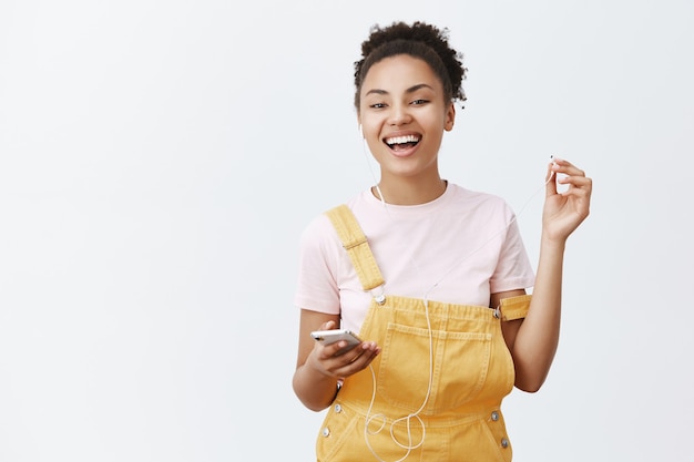 Free photo you asked something. portrait of carefree relaxed and happy african american female in trendy overalls, taking off earphone to hear friend, smiling broadly, holding smartphone, listening music