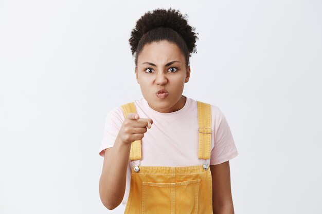 You bastard. Portrait of angry and pissed woman recognizing man who stole purse, pointing with anger and disgust, frowning, pursing lips and bending head, posing over grey wall