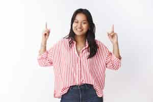 Free photo you better lift head up. portrait of enthusiastic and carefree young asian pretty girl in striped blouse smiling carefree as pointing upwards with raised hands inviting see interesting promotion