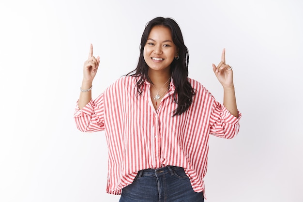 You better lift head up. Portrait of enthusiastic and carefree young asian pretty girl in striped blouse smiling carefree as pointing upwards with raised hands inviting see interesting promotion