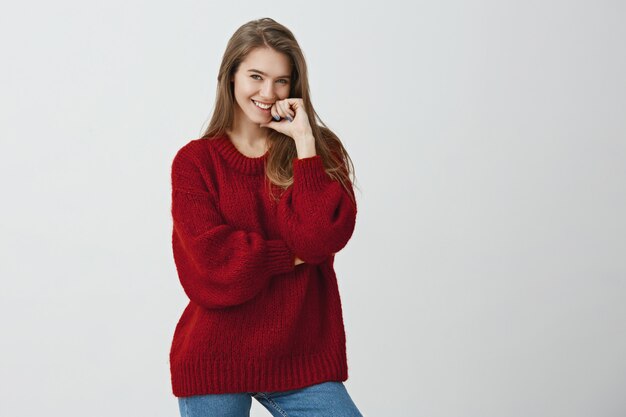 You caught my attention. Studio shot of positive attractive female coworker in trendy loose sweater holding finger on lip while smiling with interest or desire, listening to handsome guy