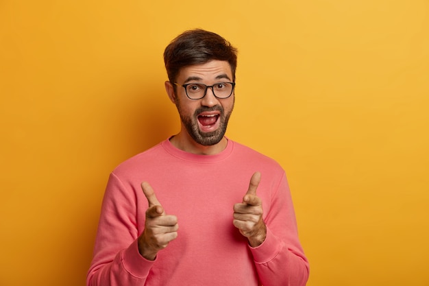 You, go with me. playful positive unshaven man welcomes friend and says hi, points with finger gun gesture, greets or congratulates person, wears casual jumper, poses over yellow wall