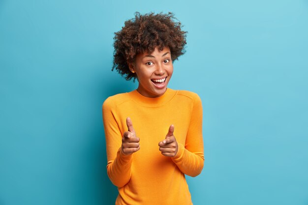 You got it. Happy young curly woman with curly hair points at camera with index fingers picks someone smiles broadly isolated over blue studio wall