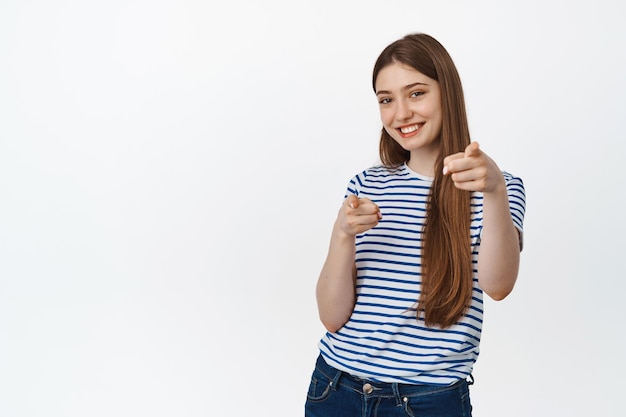 Foto gratuita l'hai capito. giovane donna sorridente che punta il dito verso la fotocamera, scegliendo, invitando le persone, in piedi in abiti casual su sfondo bianco