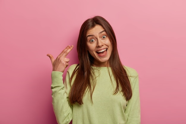 You kill my vibe. Funny dark haired woman makes gun gesture near head, shoots in temple, has glad expression, kills herself, wears green jumper