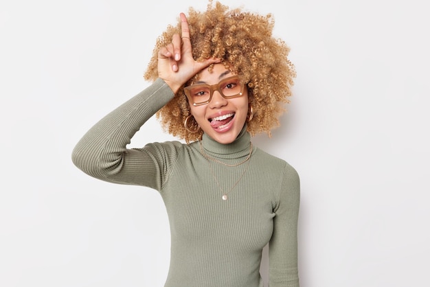 Free photo you lost funny young woman with curly bushy hair scolds friend for losing makes loser gestur over head and sticks out tongue has fun dressed in casual poloneck isolated over white background