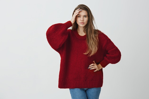 You made my head ache from boring talks. Indoor portrait of beautiful girl in winter sweater holding palm on forehead and standing with tired and exhausted expression 