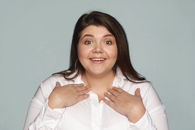 You mean me. Fascinated amazed young plus size woman in white shirt pointing at herself, holding hands on her chest and smiling broadly as being chosen for job positive among other candidates