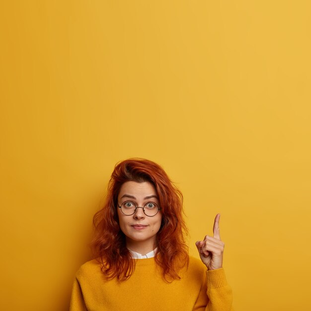 You need look. Surprised redhead European woman wears round glasses, points above, shows brand new product, advertises copy space