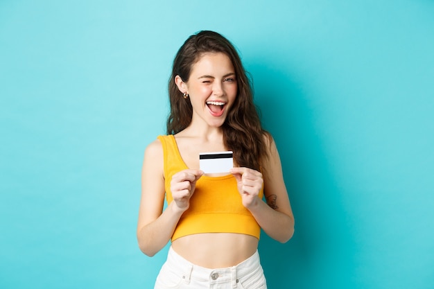 You need this. Smiling attractive woman hinting, winking at you and showing plastic credit card, recommending contactless payment, standing over blue background.