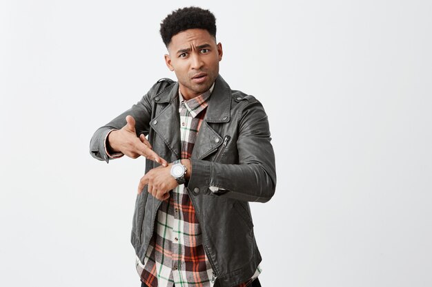 You're late. Portrait of angry beautiful black-skinned mature guy with afro hairstyle in leather jacket pointing on hand watch with unsatisfied expression after friend being late for hour.