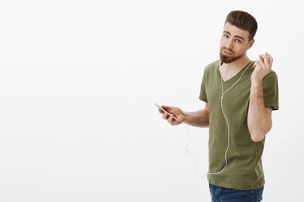 You said something. Portrait of handsome boyfriend standing in profily at subway turning and taking of earphone as asking what person wanted say holding smartphone over white wall