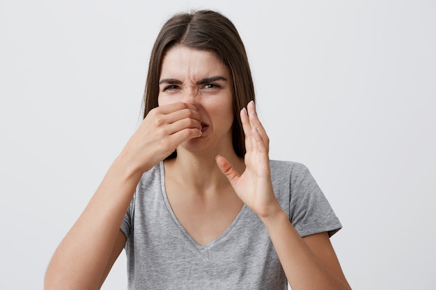 You should take a shower. Portrait of funny good-looking young caucasian woman with long dark hair in casual shirt clothing nose with fingers,  with angry expression, feeling disgust