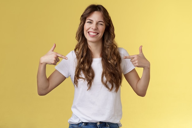 You would not regret this. Sassy good-looking outgoing daring young woman pointing center copy space indicating herself winking joyfully show perfect candidature stand yellow background
