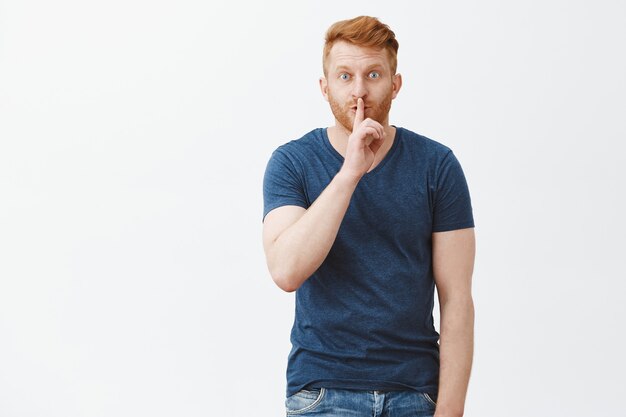 Youn cannot tel anyone, keep it secret. Portrait of charming intriguing handsome redhead with bristle, saying shh while showing shush gesture with index finger over mouth, telling rumor to friend