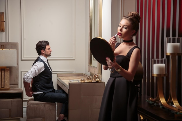 Free photo youn handsome man sitting and looking at the mirror and woman painting her lips in elegant dark rom