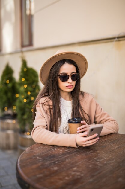 Yound fashion model lady is sitting on the table at cafe dresses in casual clothes dark sunglasses with coffee cup and phone