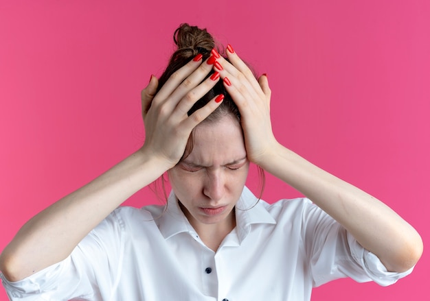 Young aching blonde russian girl holds head with closed eyes isolated on pink space with copy space